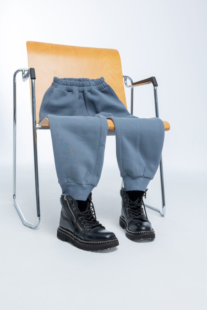 Gray sweatpants with black boots lying on wooden chair on light background. Vertical photo.