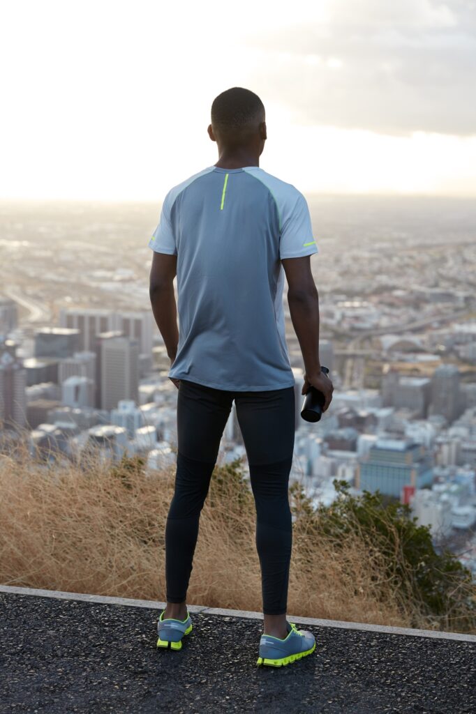 Outdoor vertical shot of athletic man wears sportclothes, stands back, admires nature view and city